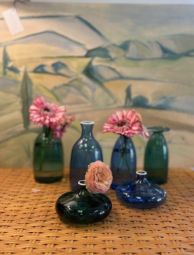 Set of glass vases with flowers on a woven surface in front of a landscape painting.