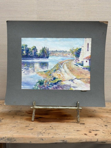 Painting of a landscape with a river and buildings on a gray mat, displayed on a wooden easel.