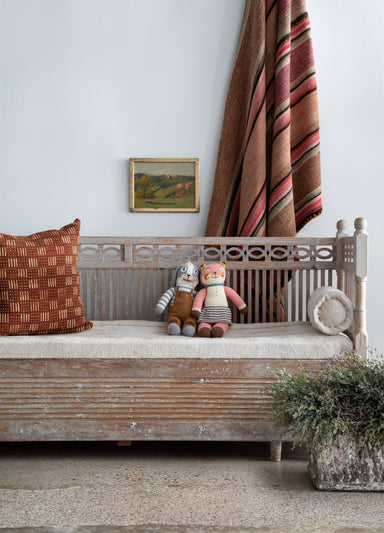 antique Swedish wood bench with upholstered seat and two bolster pillows; shown with vintage concrete planter, striped rug hung on the wall and a small vintage oil painting